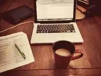 A cozy workspace setup for work from home using pay per click, with an open laptop, a notepad with a pen on top, and a warm cup of coffee, suggesting a productive session in a